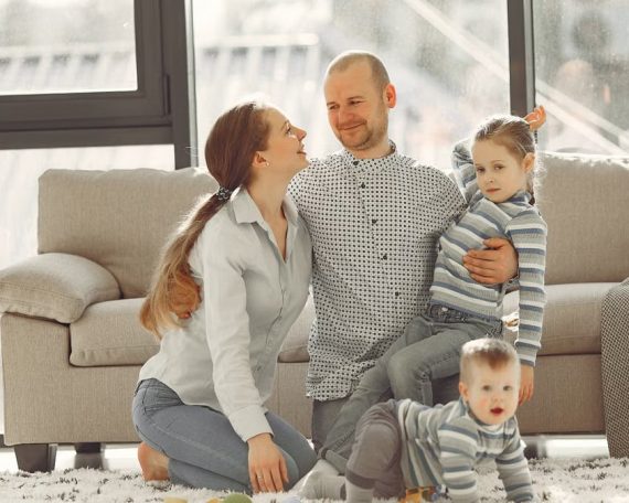 Cheerful mother and father smiling at each other while little cute boy crawling on floor and preschool girl in striped turtleneck raising arm and looking at camera